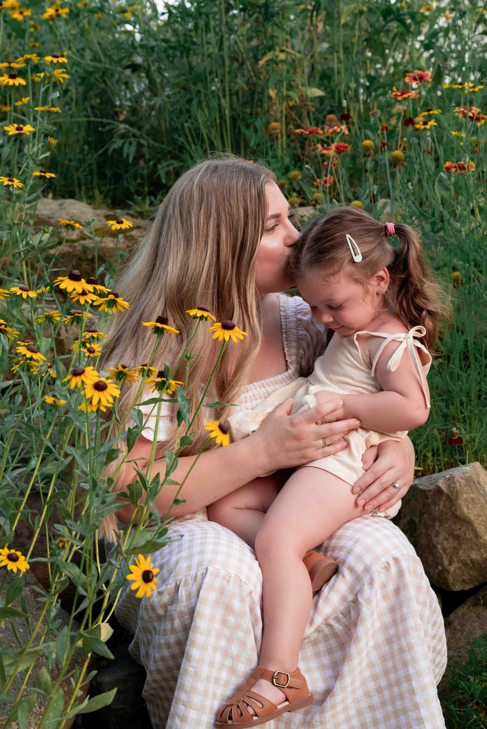 mother kisses the top of toddler daughter's head while they sit in wildflowers during a summer family photo session